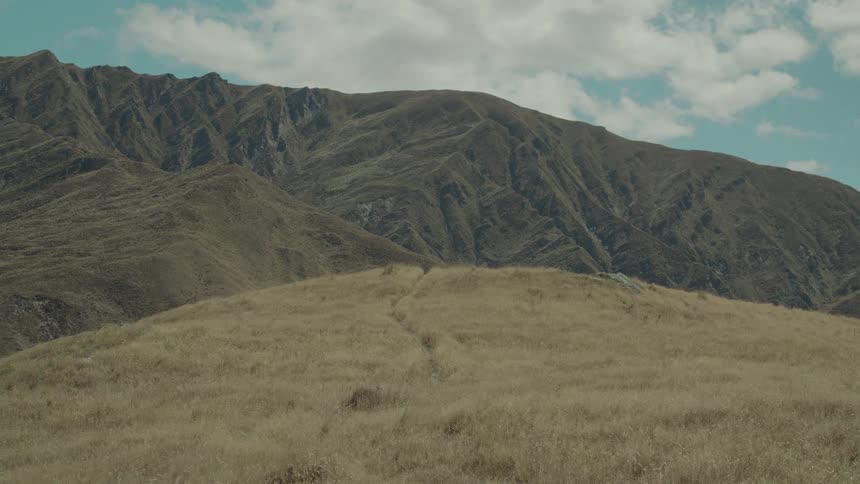 A vast landscape with a large dry grassy hill in the foreground and imposing mountains stretching across the horizon under a cloudy sky
