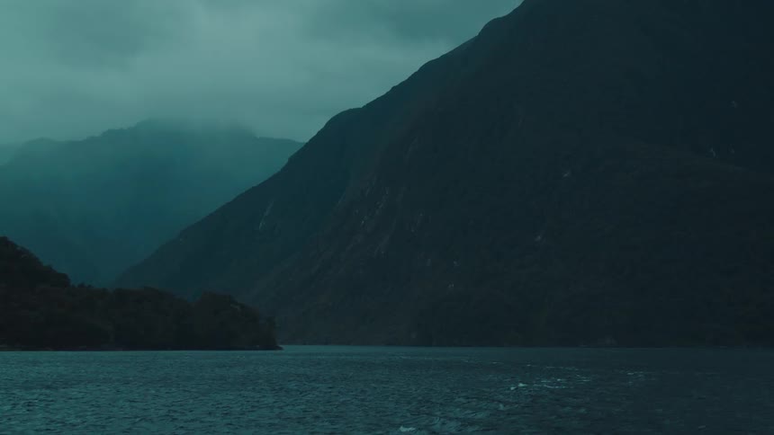 A wide dark lake rests calmly below towering misty mountains under a moody sky