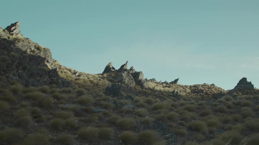 Craggy rock formations and dry yellowish grasses dominate a sunlit hillside under a clear blue sky