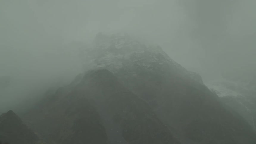 A large mountain with patches of snow visible on its upper slopes is heavily shrouded in dense fog and clouds