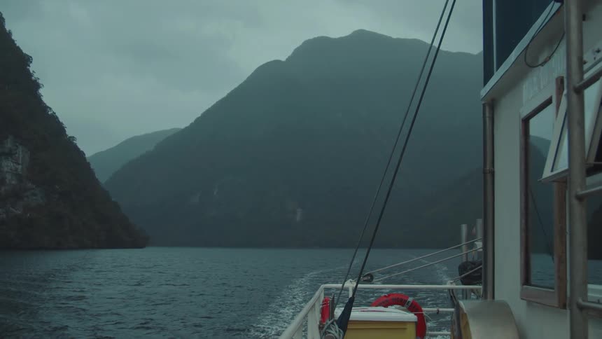 A ferry sails across a vast expanse of water framed by steep dark mountains under a cloudy sky