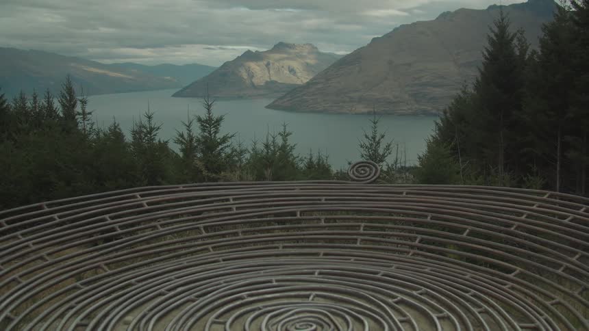 Flat dish shaped coiled sculpture occupies an area among conifers on the sloping coast across the lake from the mountains on an overcast day