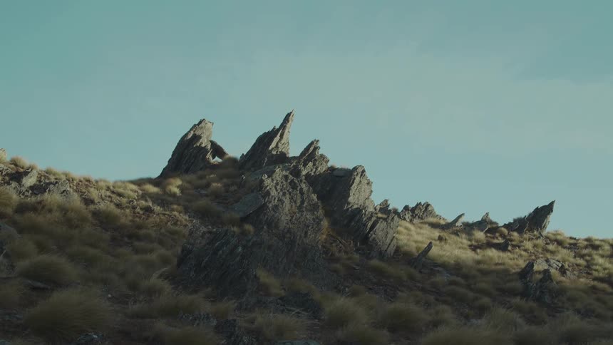 Wind ruffles the vegetation near jagged rock formations on a slope under a blue sky