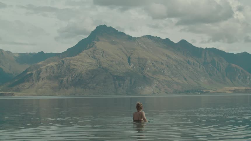 A woman in a swimsuit stands in a calm lake framed by a rugged mountain range beneath a cloudy sky