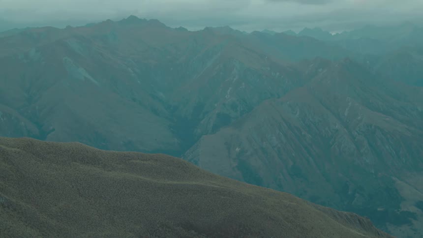 Rolling mountain ranges stretch across the horizon under an overcast sky with a dry scrubby hillside in the foreground