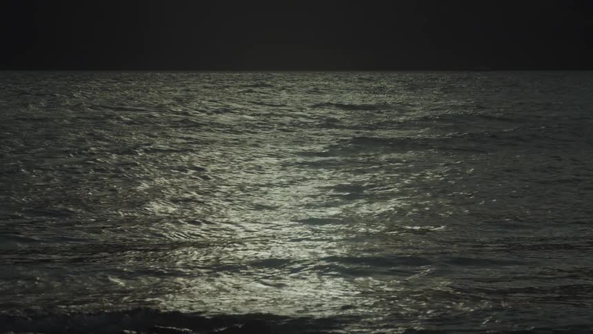 Waves break on a dark rocky shore under a dark sky with the moons reflection shimmering on the waters surface