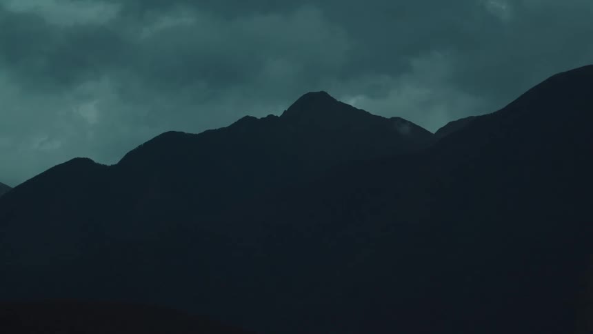 A dark silhouette of a mountain range is visible against a moody cloudfilled sky in very low light conditions
