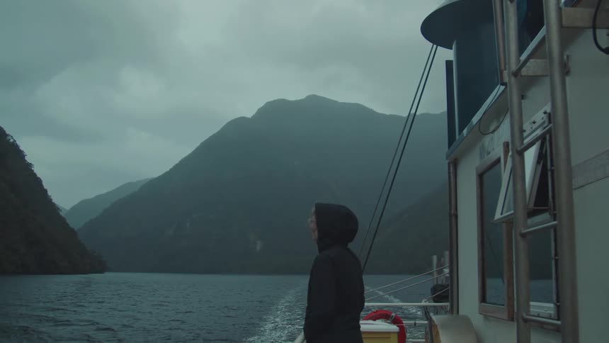 A woman on the outside of a boat looking at the scenery with calm sea and large mountains around her