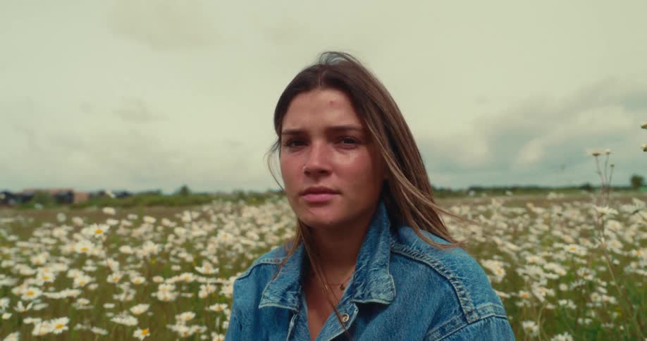 A young woman sits in a daisy field calmly looking at the camera