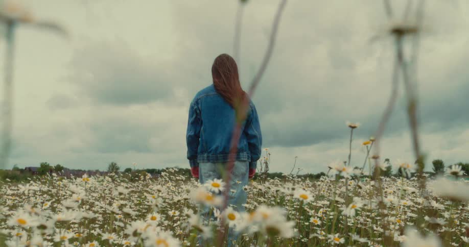 The back of a young woman stands in a field of white daisies under a cloudy sky