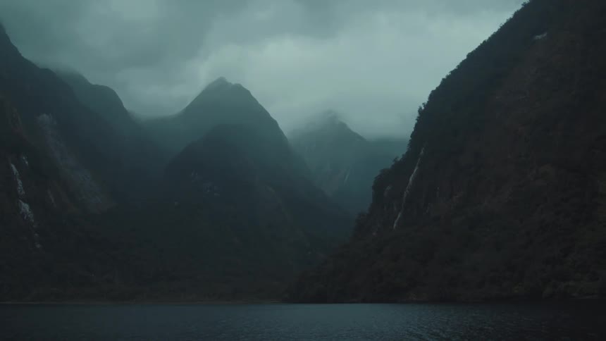 Steep mistshrouded mountains rise dramatically from the dark waters of a fjord under a moody overcast sky
