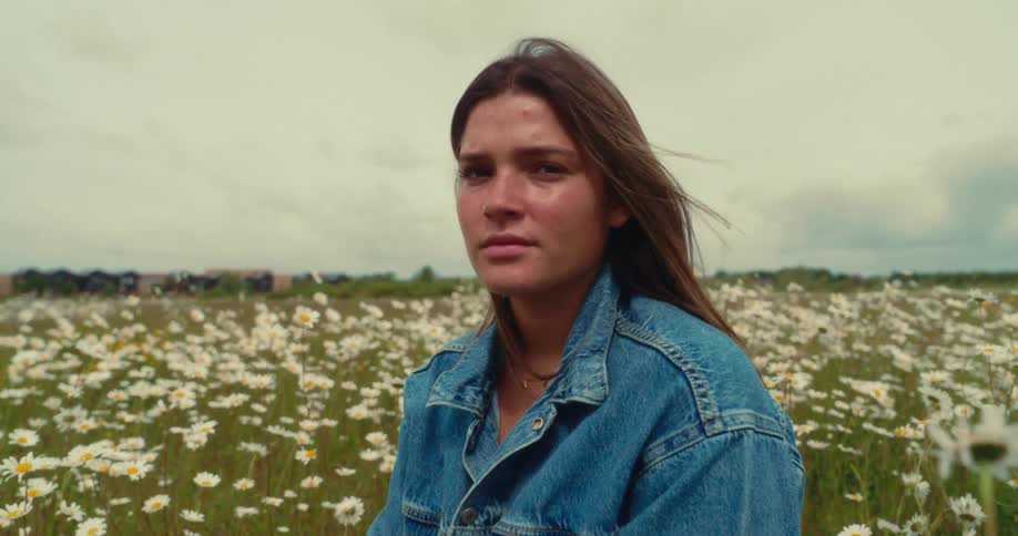 A young woman gazes calmly at the camera while sitting in a daisyfilled field
