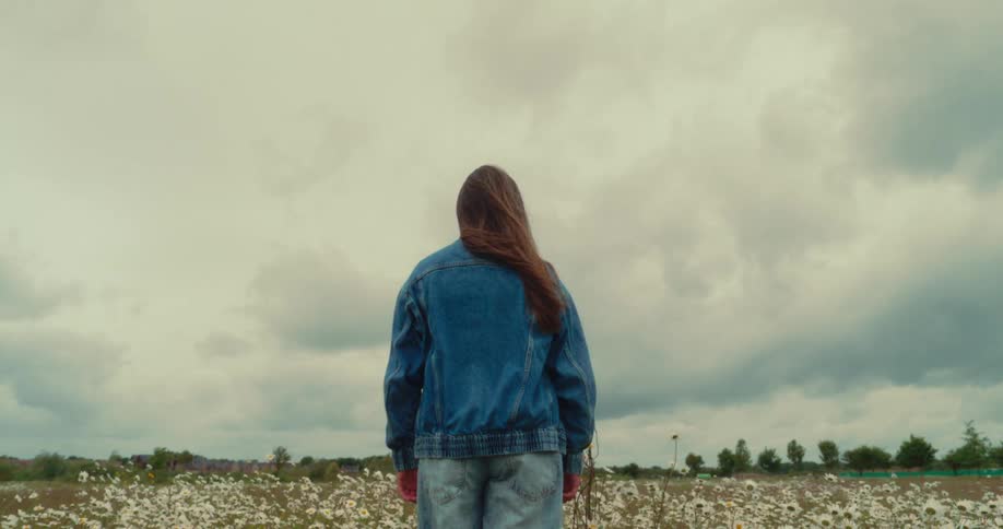 A girl stands alone in a flower field under a moody sky facing away from the camera