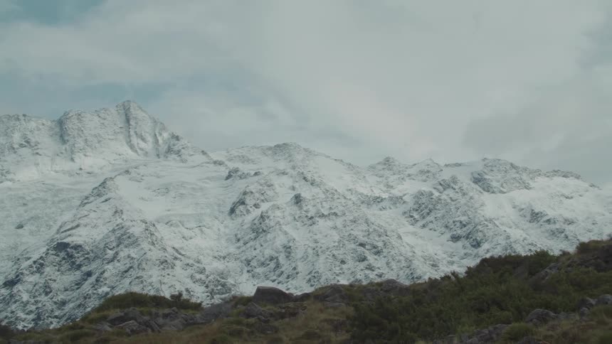 Snow covers the peaks of towering mountains under a partly cloudy light blue sky