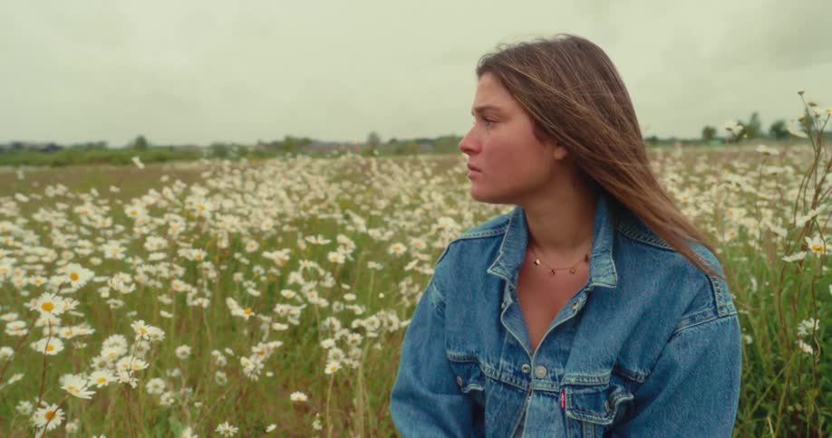 A young woman wearing a denim jacket looks off to the side in a field filled with white daisies under a cloudy sky