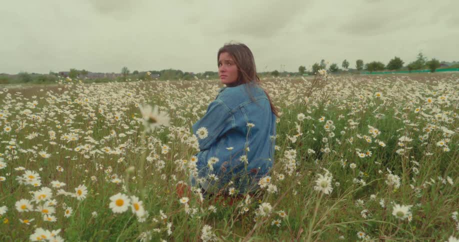 A young woman in a denim jacket turns back while sitting in a vast field of daisies