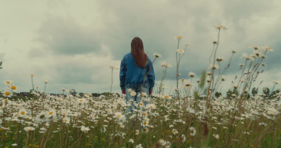 Young woman stands among wildflowers in a meadow and faces the sky during daytime