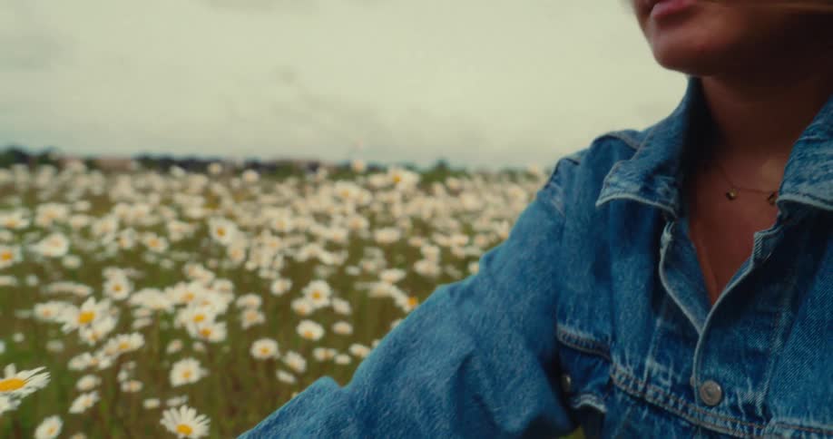 A young woman sits in a field of white daisies gently touching the flowers as the wind lifts her flowing hair