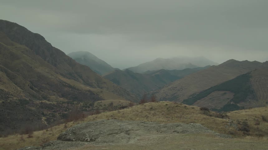 A cloudy day over some rugged mountains with a valley winding through them and some dry grassy slopes in the foreground