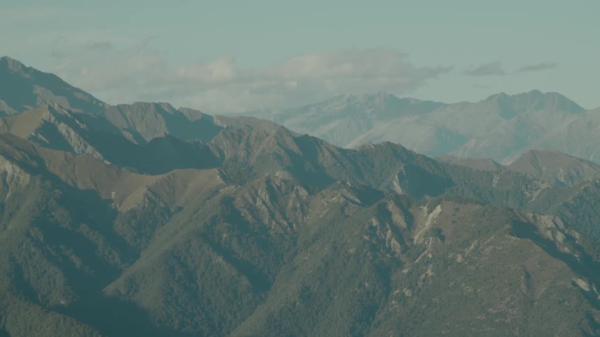 An expansive aerial view captures a rugged mountain range its slopes largely covered in vegetation and carved by deep valleys stretching towards a hazy sky