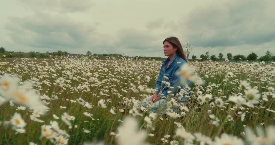A woman wearing a denim jacket is kneeling in a field of white daisies with a cloudy sky above