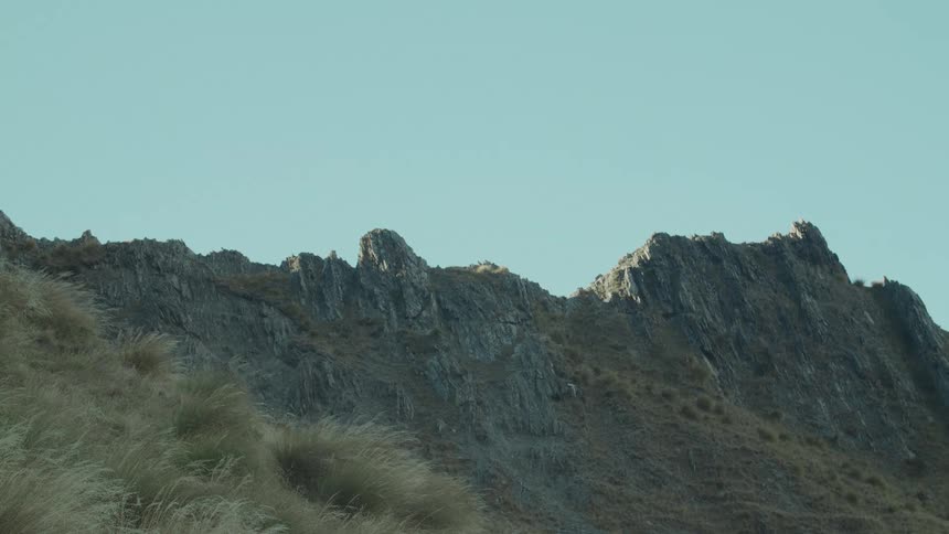 Craggy rock formations and dry yellowish grasses dominate a sunlit hillside under a clear blue sky