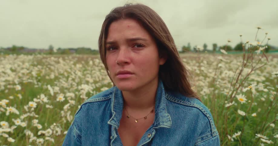 A young woman in a denim jacket sits in a daisy field looking thoughtful