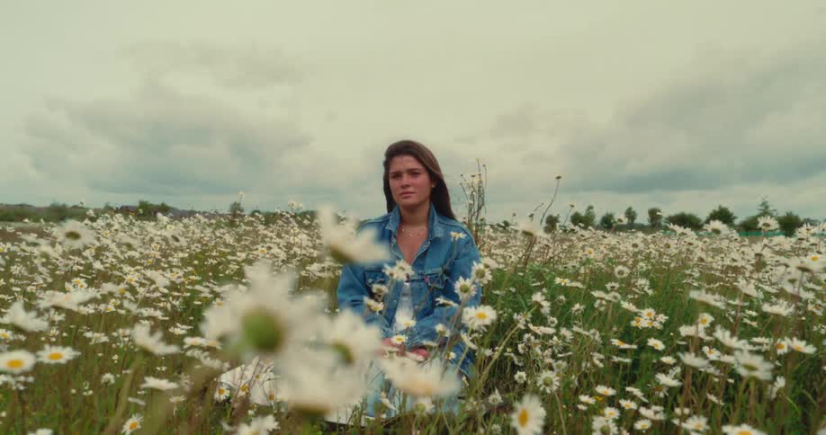 A young woman sits in a field of white daisies under a cloudy sky as the wind lifts her hair