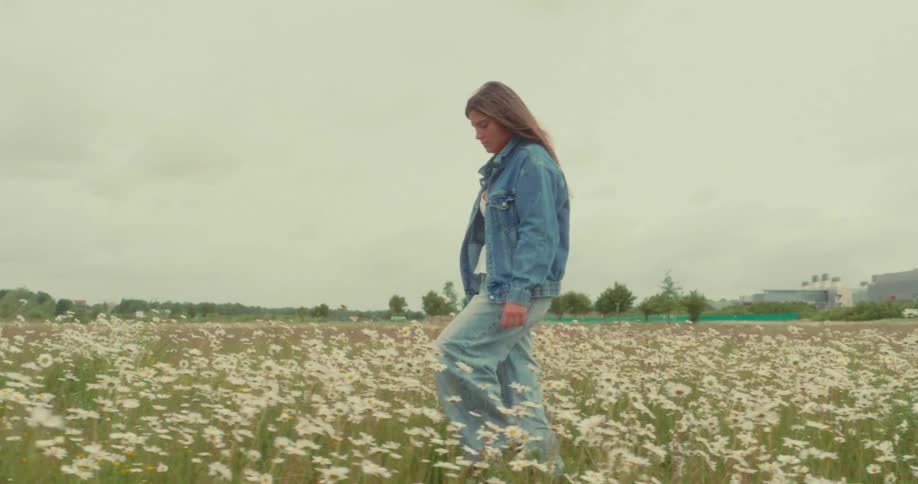 A young woman wearing a denim jacket and wideleg jeans is walking through a field full of white daisies under a cloudy sky
