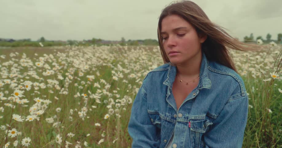A young woman sits in a field of white daisies under a cloudy sky eyes closed as the wind lifts her hair
