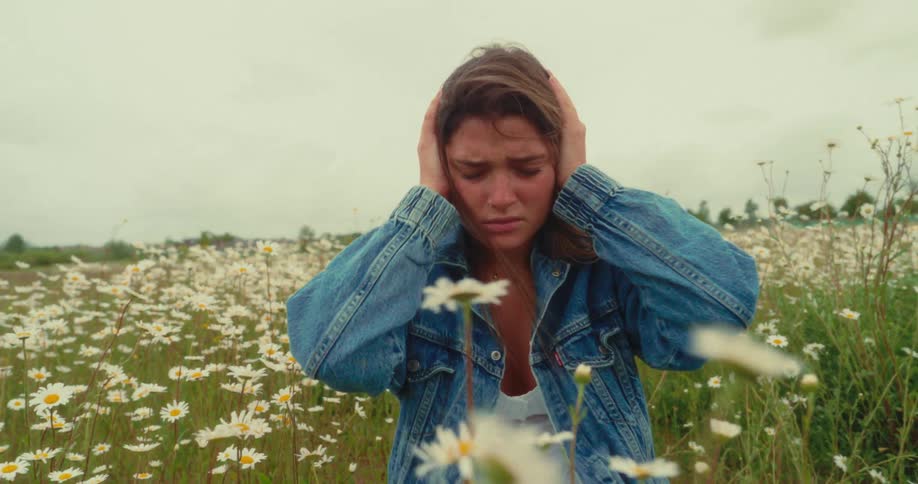 A young woman is kneeling in a field of white daisies with her hands covering her ears and an expression of distress under a cloudy sky