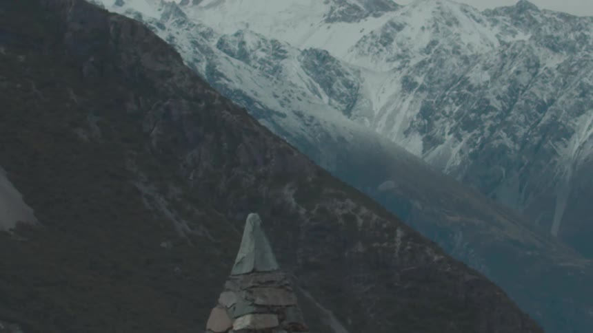 A stone cairn stands in the foreground of a vast mountain range with dramatic snowcovered peaks under an overcast sky