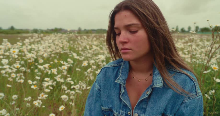 A young woman sits in a field of white daisies under a cloudy sky eyes closed as the wind lifts her hair