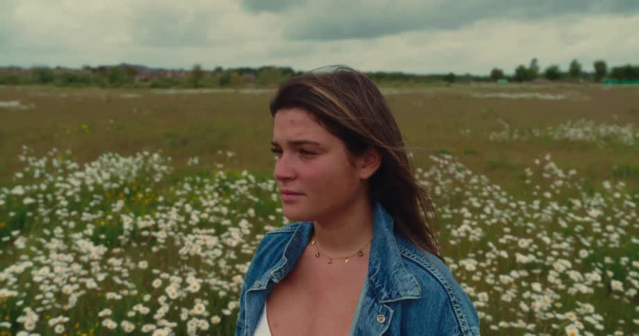 A woman in a blue denim jacket is standing in a field of white flowers and is staring straight ahead