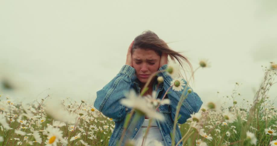 A distressed young woman is kneeling in a field of white daisies holding her hands over her ears under an overcast sky