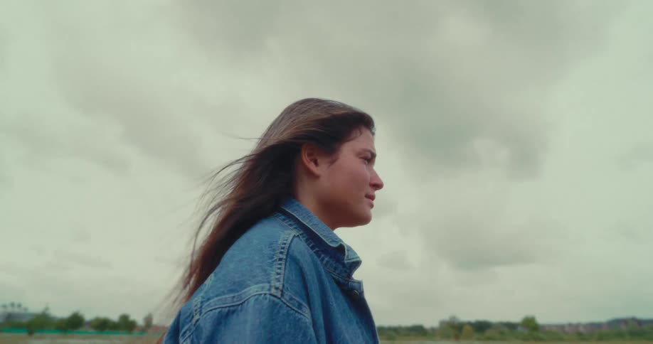 A woman in a denim jacket walks through an open field under a cloudy sky