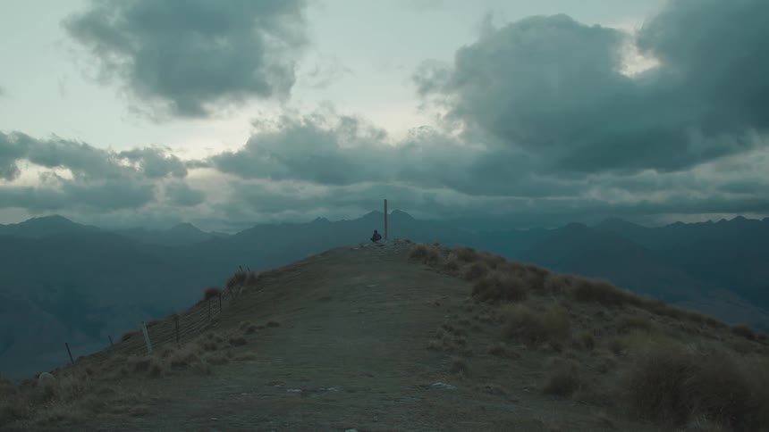 A lone figure sits atop a mountain ridge surrounded by dramatic clouds and distant peaks