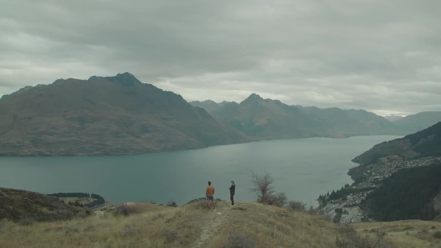 Two people standing at the edge of a cliff overlooking a lake surrounded by mountains on a cloudy day