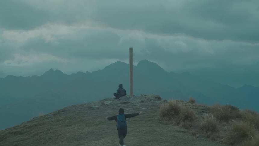 A person running toward another individual who is sitting on the edge of a cliff on a cloudy day