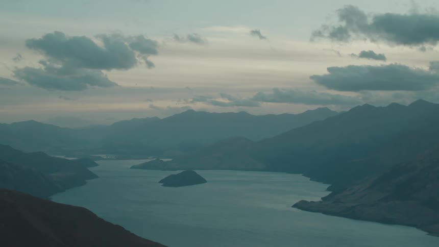 A wide dim view from a high vantage point shows a lake with a central island nestled within a valley surrounded by mountains under a cloudy sky