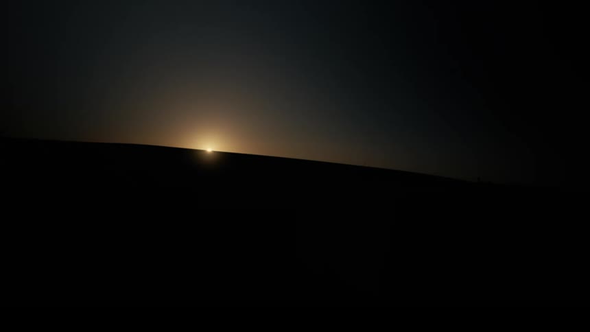 A sliver of bright light appears on the dark horizon silhouetting a hill as the sun begins to rise or set