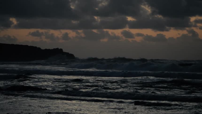 Dark slowmotion waves crash onto a beach against a silhouetted coastline under a cloudy sky at dusk or dawn