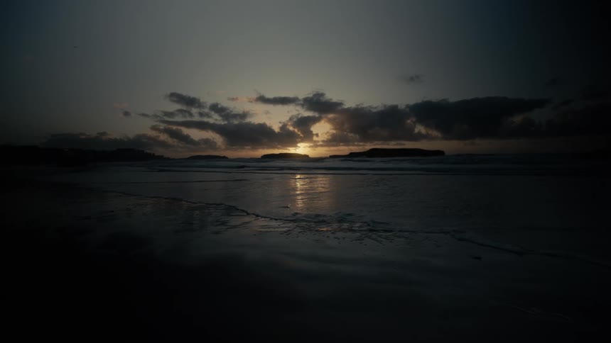 Sun sets behind a dark island silhouette casting reflections on a quiet wet beach shoreline