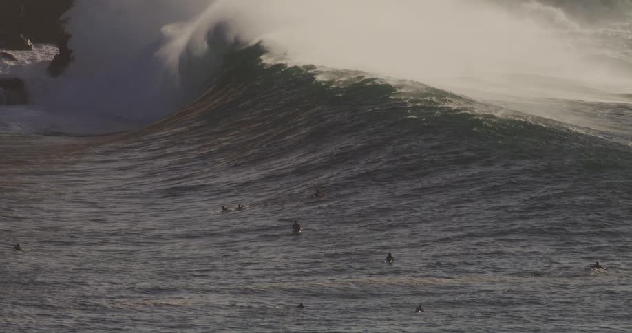 A group of people swimming and paddling on surfboards in the ocean as large waves form under the midday sun
