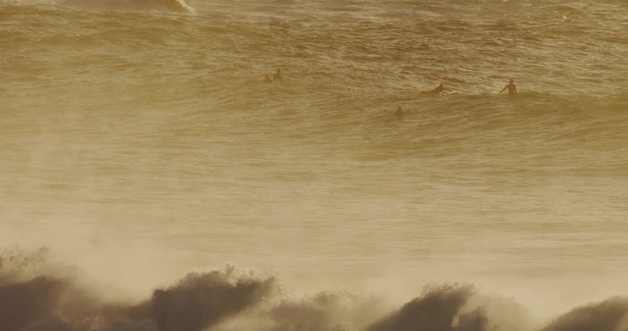 Several surfers paddle and wait in the golden turbulent ocean obscured by mist and large waves breaking in the foreground under a hazy sky