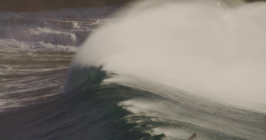 A lone surfer rides a large dark wave as ocean water cascades down jagged rocky cliffs in the background