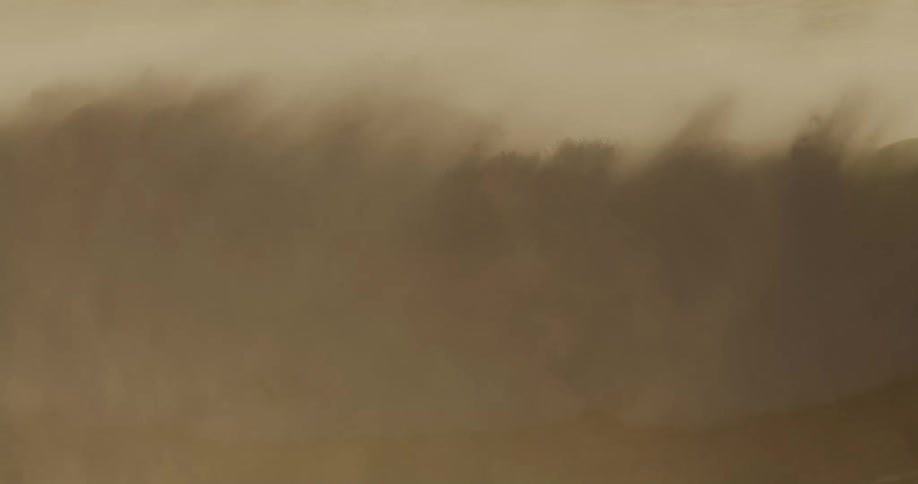 A surfer riding a monstrous ocean wave as mist rises from the crashing water creating a foggy atmosphere at midday