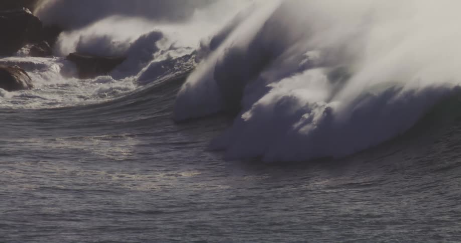 Large white waves crash powerfully against dark rocky shore on with surfers waiting