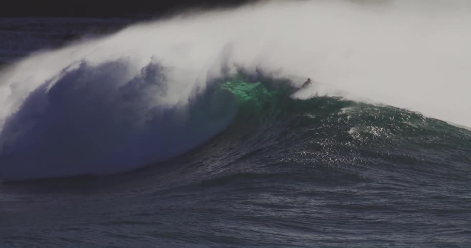 A group of surfers in the ocean ride over forming waves creating large foamy splashes under the bright midday sun