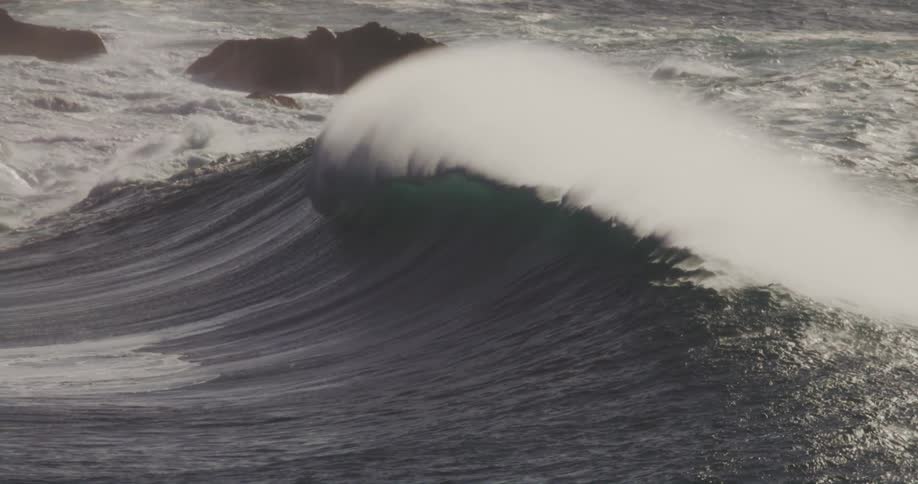 A large ocean wave crests and crashes generating a massive plume of white spray as several surfers paddle in the foreground and rocky formations appear in the background
